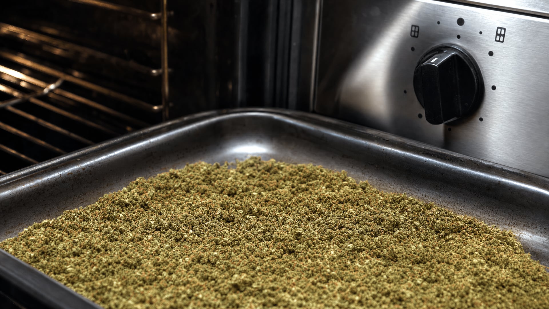 Ground cannabis on a baking tray beside an oven temperature control in a kitchen